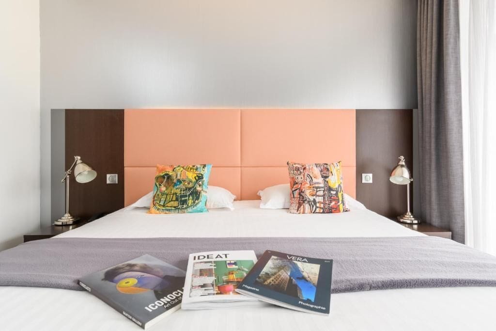 Habitación de hotel con cama, dos almohadas, una revista y una lámpara sobre una mesita de noche.