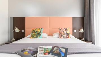 Habitación de hotel con cama, dos almohadas, una revista y una lámpara sobre una mesita de noche.