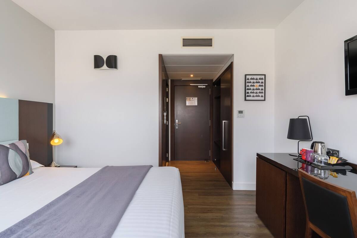 A hotel room with a bed, bedside lamp, desk with a lamp, and a door leading to another room.