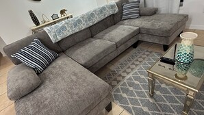Ein graues Ecksofa mit gestreiften und einfarbigen Kissen, einer gemusterten Decke, einem Glastisch mit einer Vase und einem geometrischen Teppich.
