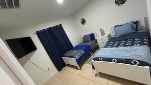 Ein kleines, gut beleuchtetes Schlafzimmer mit einem Einzelbett, einem Nachttisch, einem Wand-Fernseher und einem Fenster mit Vorhängen.