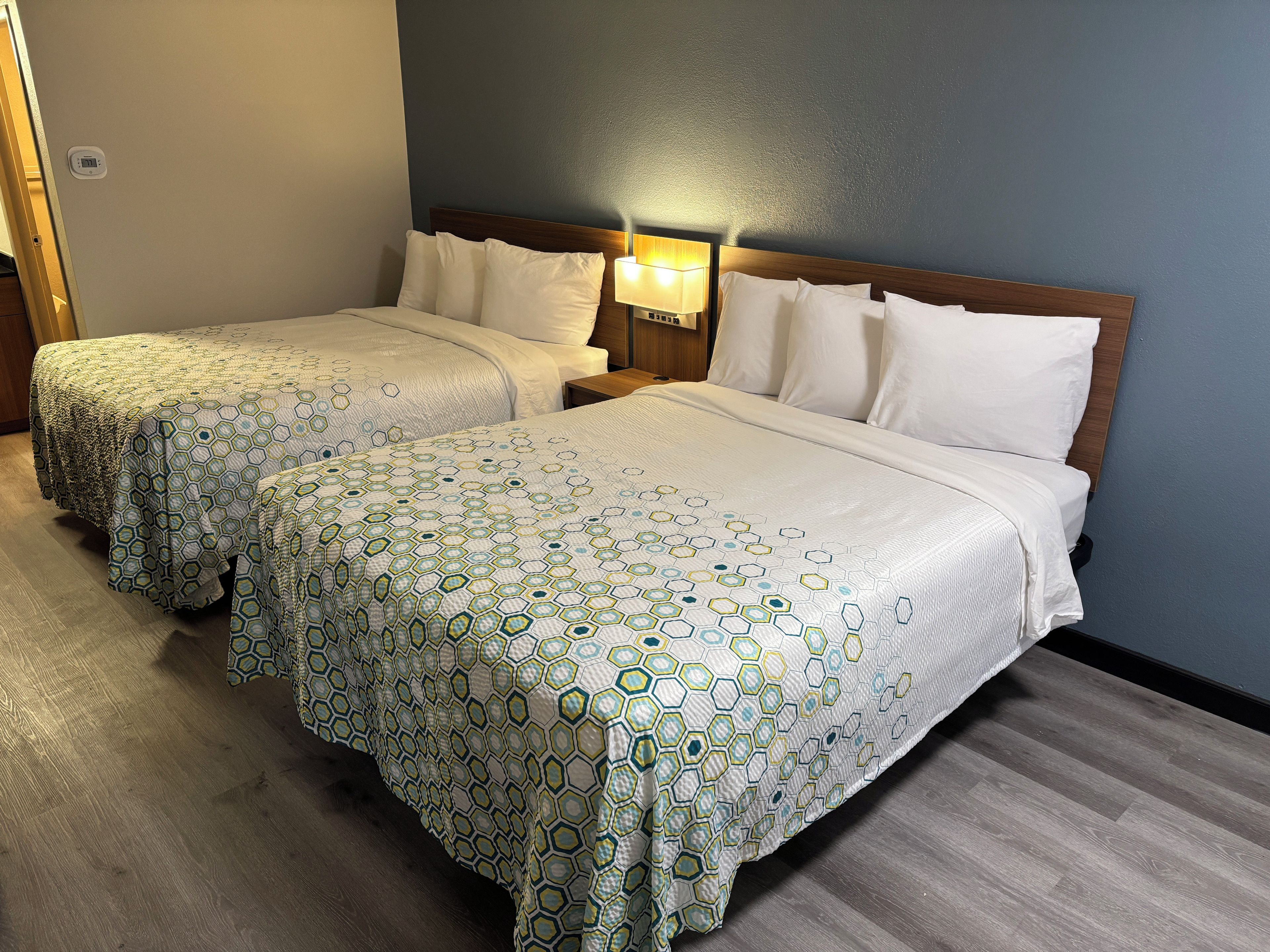 Une chambre d’hôtel avec deux lits, chacun recouvert d’une couvre-lit à motifs, une table de chevet avec une lampe et un thermostat fixé au mur.