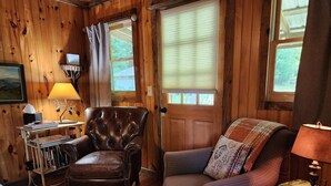 L’intérieur chaleureux d’un chalet avec un fauteuil en cuir, un fauteuil rembourré, une petite table en bois avec une lampe et une bibliothèque.