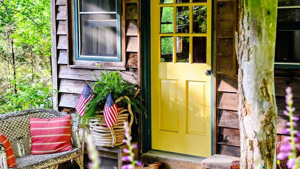Une cabane rustique avec une porte jaune, une véranda avec un fauteuil en osier et un drapeau américain.