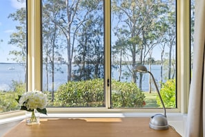 A room with a large window overlooking a body of water and trees. There is a desk with a lamp and a vase of flowers. - Waterfront Paradise Lodge Brightwaters (Brightwaters)