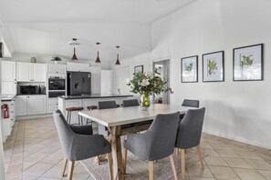 A modern kitchen with white cabinets, a central island, and a dining area with a white table and grey chairs.