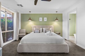 A bedroom with a large bed, two bedside tables, a chair, a ceiling fan, and a view of the outdoors.