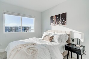 A bedroom with a large window, a bed with white bedding, a bedside table with a lamp, and artwork on the wall. - NEW Mountain Game Room Getaway (Bozeman)