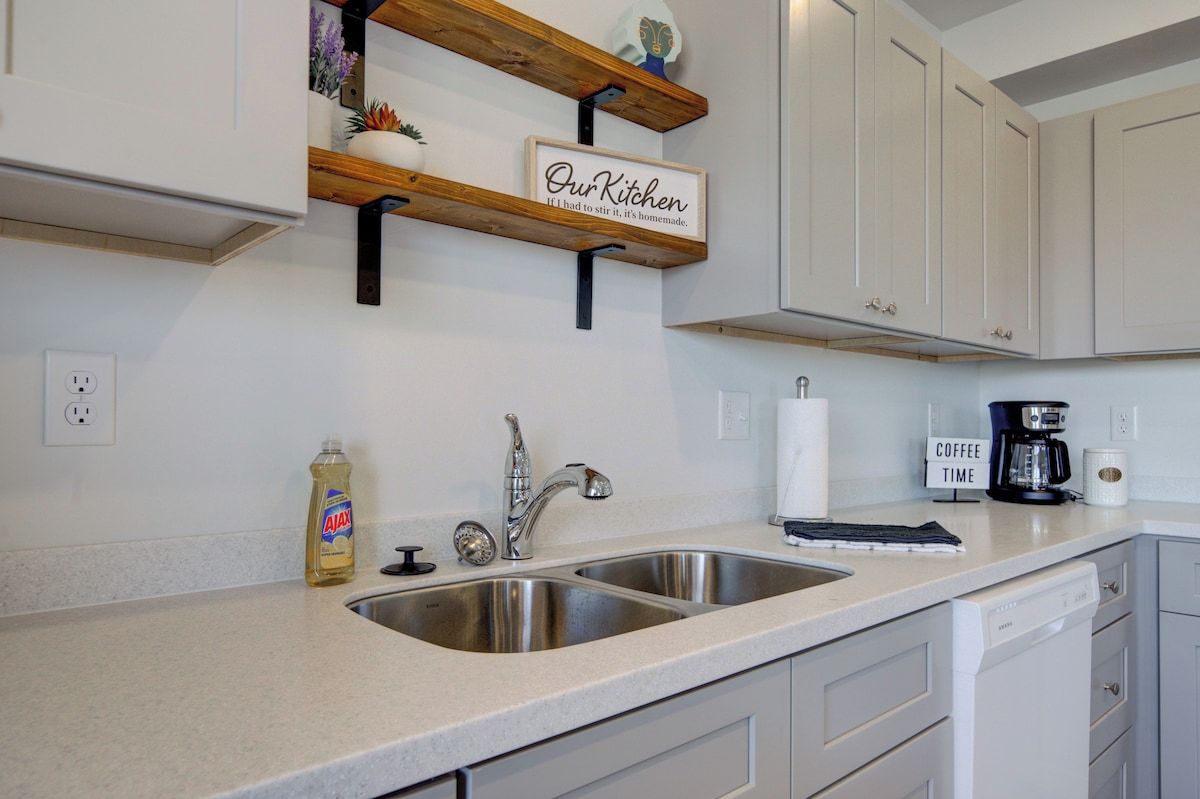 A kitchen with white cabinets, a stainless steel sink, a coffee maker, and a sign that reads 'Our Kitchen.'