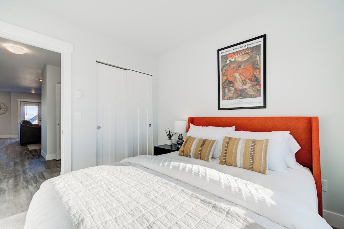 A bedroom with a bed, a framed movie poster, a white door, and a hallway leading to another room.