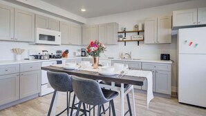 Uma cozinha moderna com armários brancos, uma ilha central com banquetas, micro-ondas e geladeira.