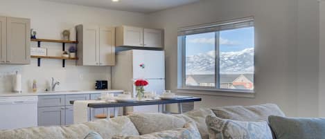 A modern kitchen with white cabinets, a dining table with a vase of red flowers, and a view of snow-capped mountains through a large window.