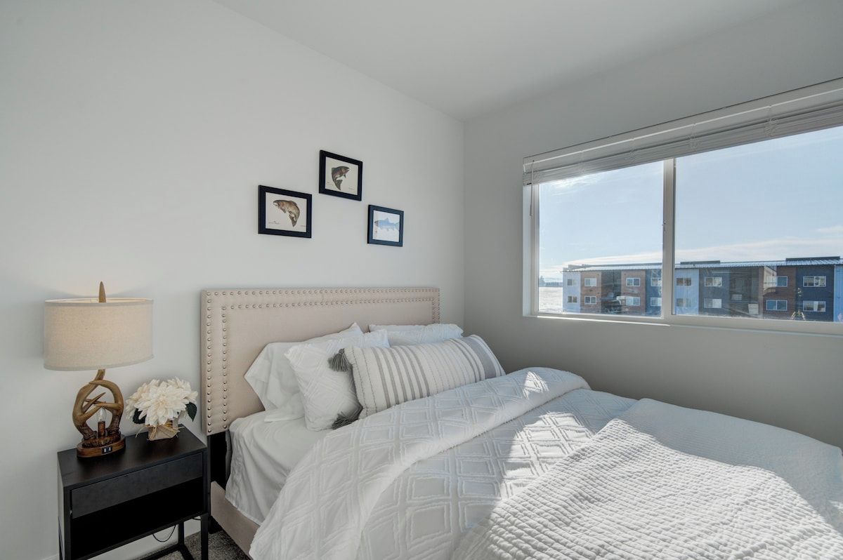 Un dormitorio con cama, mesita de noche, lámpara y ventana con vistas a edificios.
