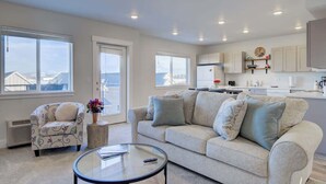 A spacious living room with a sofa, armchair, and coffee table. A kitchen with white cabinets and a dining area is visible in the background. - NEW Mountain Views | Hot Springs | Truck Stop (Bozeman)