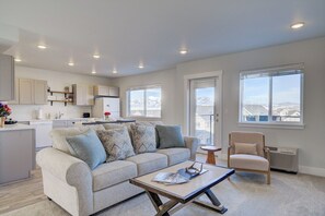 A modern living room with a sofa, coffee table, and a chair. There is a kitchen area with cabinets and a refrigerator. Large windows provide a view of the mountains.