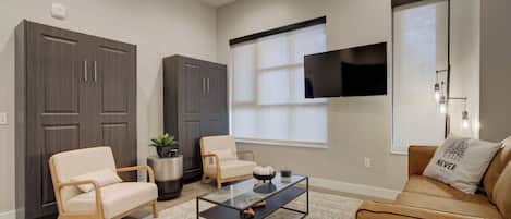 A living room with a sofa, two armchairs, a coffee table, a TV mounted on the wall, and a large window.