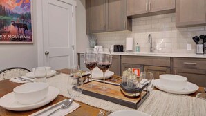 A dining area with a wooden table set for a meal, a painting of a mountain landscape, and a kitchen with modern appliances and cabinets. - Cozy Downtown Condo (Bozeman)