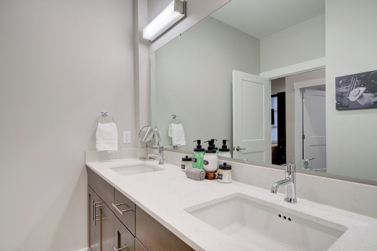 A modern bathroom with two sinks, a large mirror, and a door leading to another room.