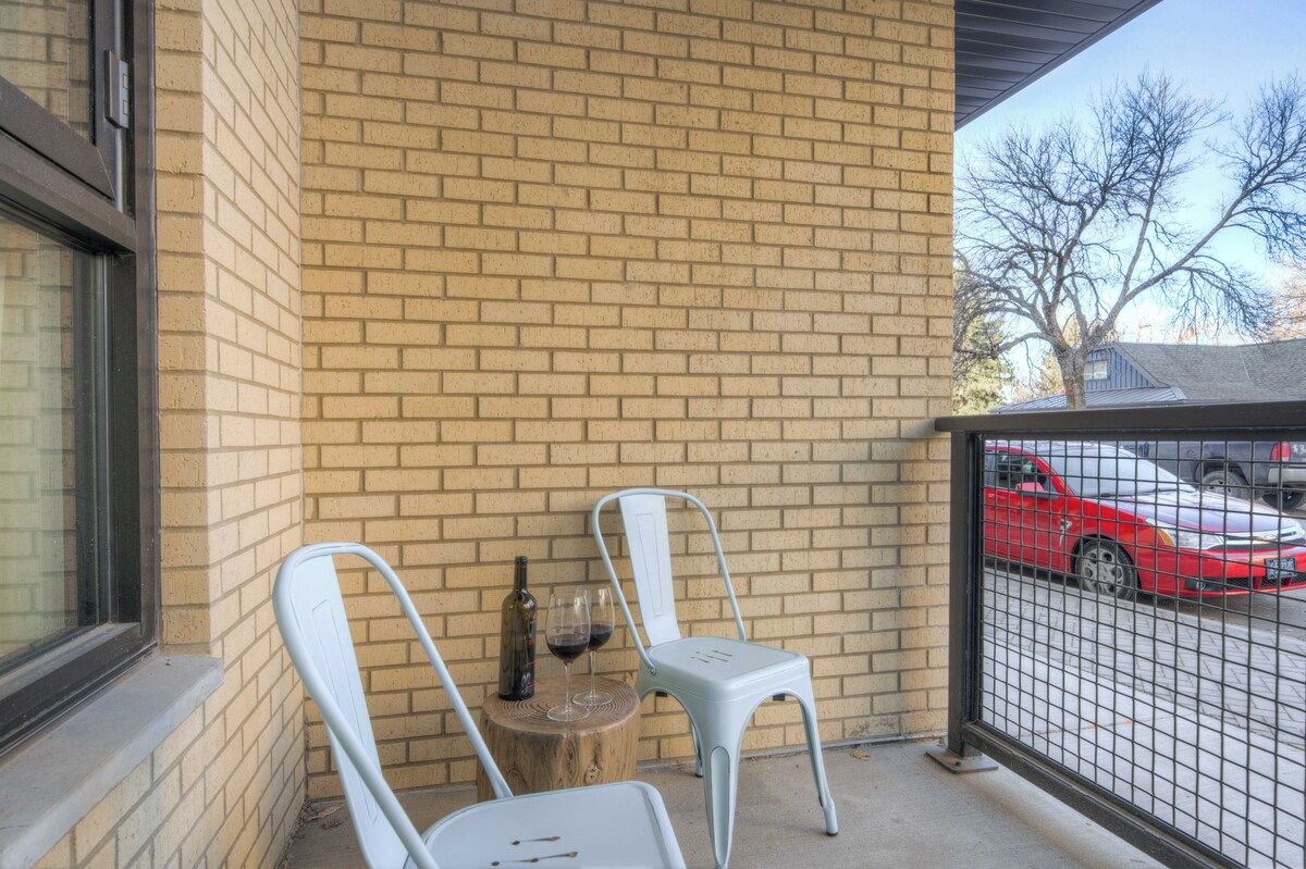 A brick wall with two white chairs and a bottle of wine on a wooden stool.