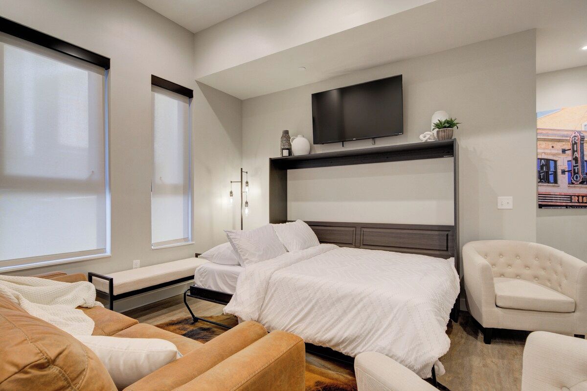 Un dormitorio moderno con una cama grande, un televisor montado encima y una zona de estar con un sofá y sillones.