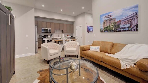 A modern living room with a glass coffee table, beige sofa, and a painting of a building on the wall. - Downtown Luxury Loft | Extreme Walkability (Bozeman)