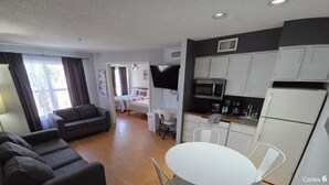 A compact living space with a sofa, a dining table, a kitchenette, and a bedroom with a bed and a TV.