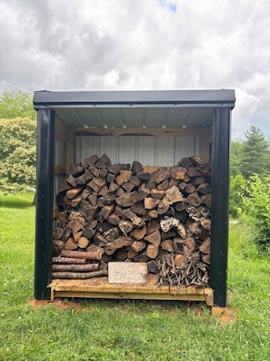 A small, dark-colored shed with a metal roof, filled with neatly stacked firewood and a few branches on the floor.