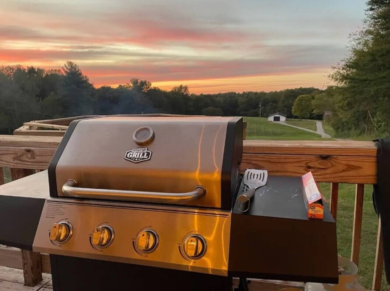 A stainless steel grill with four control knobs and a side burner, placed on a wooden deck with a scenic view of trees and a sunset in the background.