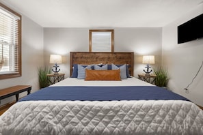 A bedroom with a large bed, two bedside tables with lamps, a wooden headboard, a window with blinds, and a wall-mounted TV.