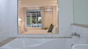 A modern bathroom with a large bathtub, a glass-enclosed shower area, and a view of a bedroom with a bed and curtains. - Squirrel House (Bangkok)