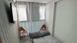 A compact room with a grey sofa, two wooden chairs, a small table, and a ceiling-mounted air conditioner. - Flats a 450m do mar em Porto de Galinhas (Ipojuca)