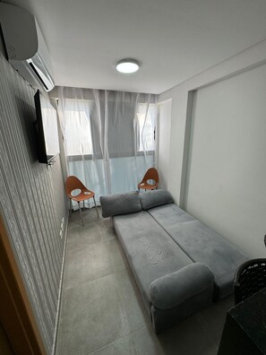 Een compacte kamer met een grijze bank, twee houten stoelen, een klein tafeltje en een luchtverwarmer aan het plafond.