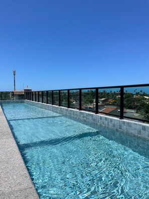 A clear blue swimming pool with a tiled edge, surrounded by a black railing, and a view of residential buildings and a tall communication tower in the background.