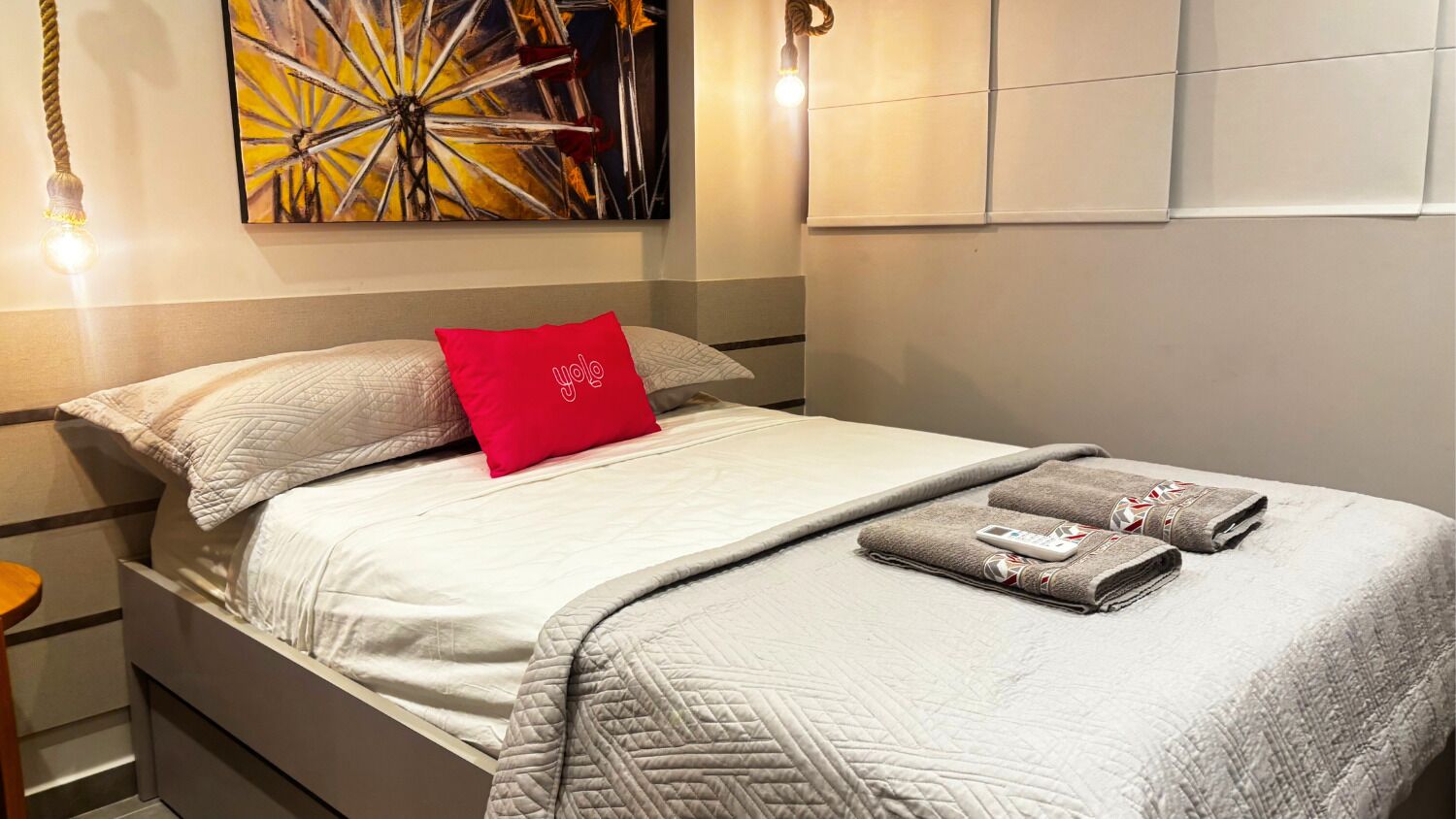 A neatly made bed with a red pillow, a folded towel, and a remote control on it.