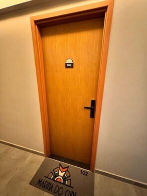 A wooden door with a number plate '102' and a doormat with the text 'MARINA DO CUDE'.