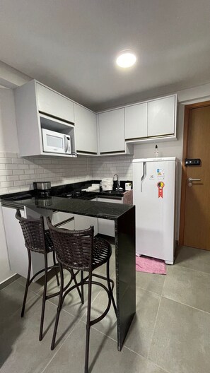 Una cocina compacta con armarios blancos, un microondas, un refrigerador y dos sillas de mimbre.