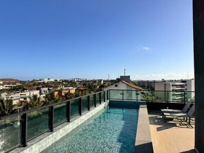 Una piscina en la azotea con agua cristalina y azul, rodeada por una barandilla de vidrio, con vistas a un paisaje urbano con edificios y palmeras.