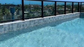 A clear blue pool with a tiled edge, surrounded by a glass railing, overlooking a residential area with houses and palm trees. - Flats a 450m do mar em Porto de Galinhas (Ipojuca)