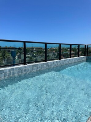 Een helderblauw zwembad met betegelde rand, omgeven door een glazen balustrade, met uitzicht op een woongebied met huizen en palmbomen.