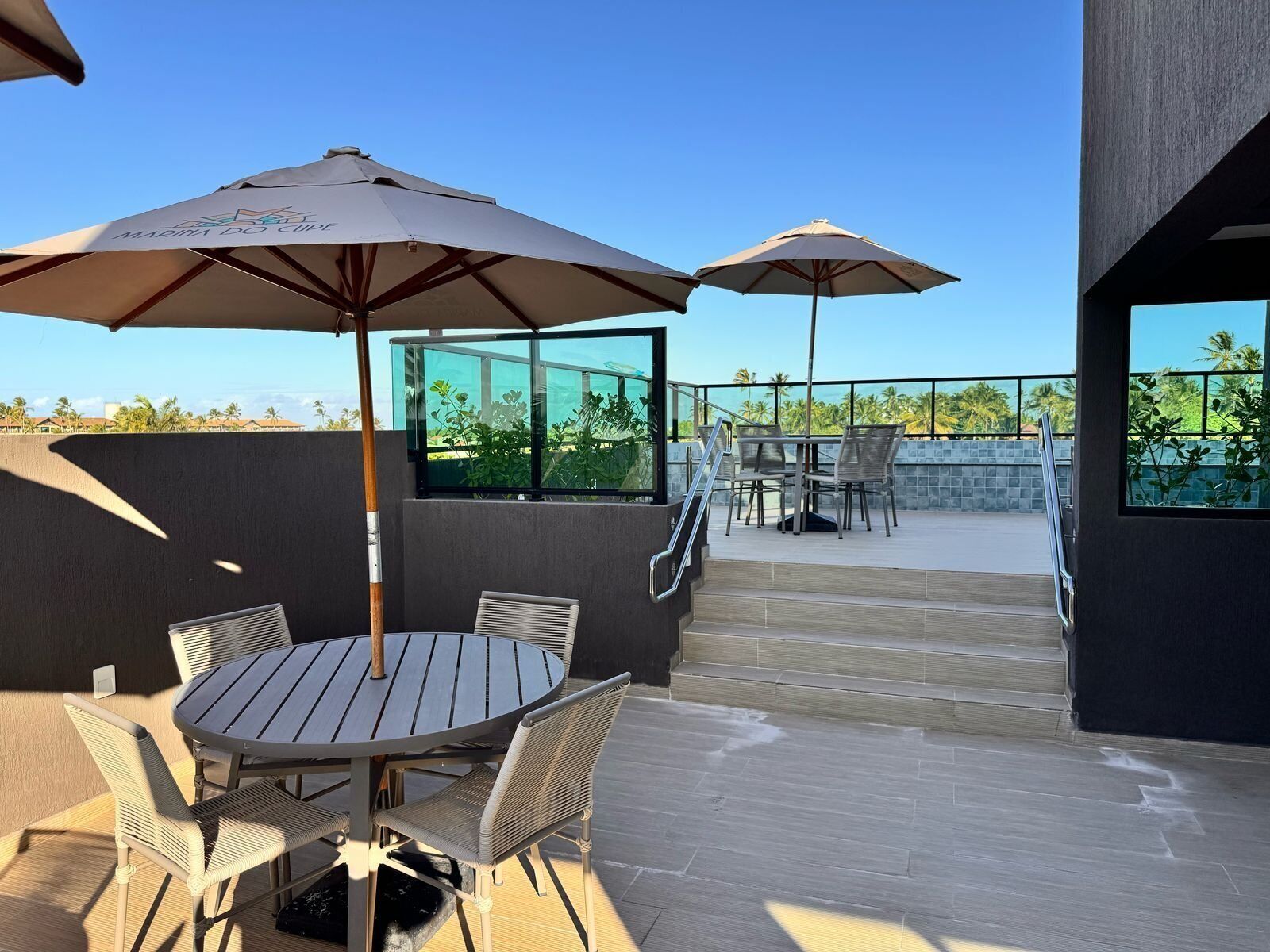 An outdoor patio with a round table and chairs, two umbrellas, and a view of palm trees.
