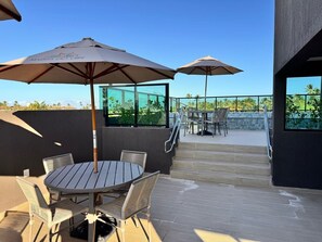 En udendørs terrasse med et rundt bord og stole, to parasoller og udsigt til palmer.