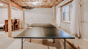 Una mesa de ping pong en una habitación con vigas de madera a la vista, una ventana con cortinas y una mesa con sillas.