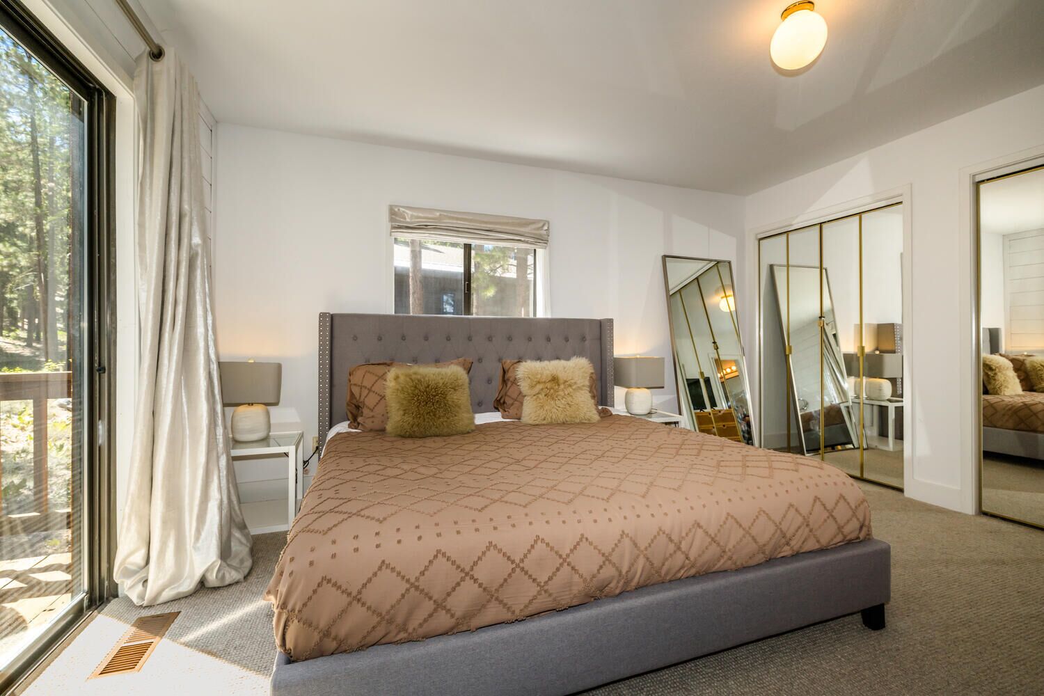 A modern bedroom with a large bed, a window with curtains, a bedside table with a lamp, and a mirror on the wall.