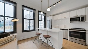 Una cocina moderna con armarios blancos, una isla central con taburetes, electrodomésticos de acero inoxidable y grandes ventanales con vistas a la ciudad.