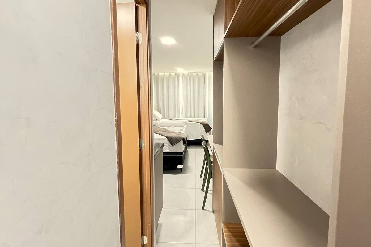 A narrow hallway with a wooden door on the left, a bed with white bedding in the background, and a green chair.