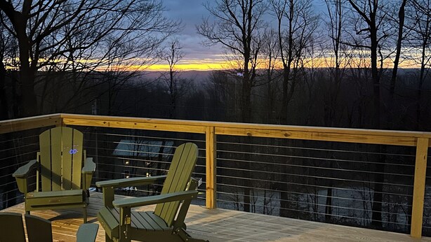 A wooden deck with Adirondack chairs and a table, overlooking a forested area with a view of the sunset.