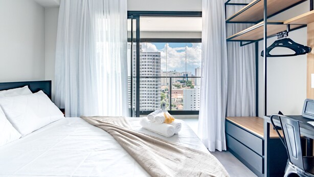 A hotel room with a bed, a desk, and a view of buildings through large windows. - Refuge in S o Paulo With Swimming Pool Sp-p2003b (São Paulo)