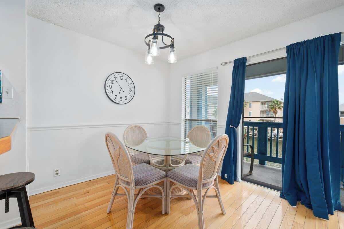 Een eetgedeelte met een ronde glazen tafel, rieten stoelen, een wandklok en uitzicht door een balkon met blauwe gordijnen.
