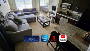 A living room with a sofa, a coffee table, and a TV. There's a kitchen with a microwave and cabinets.