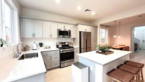 A modern kitchen with stainless steel appliances, white cabinetry, and a central island with stools. - New! 4BR Vacation Home, Tub, BBQ, EV, close to Sequoia (Visalia)